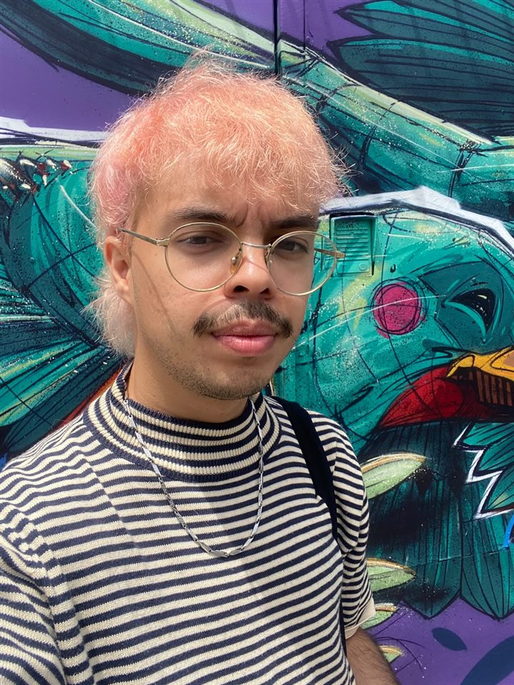 Adrián Betancourt, with pink hair and round glasses, in front of a colorful mural